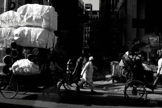 A man, a rickshaw and a mountain of barrels crossing the busy streets of Dhaka amidst the traffic.